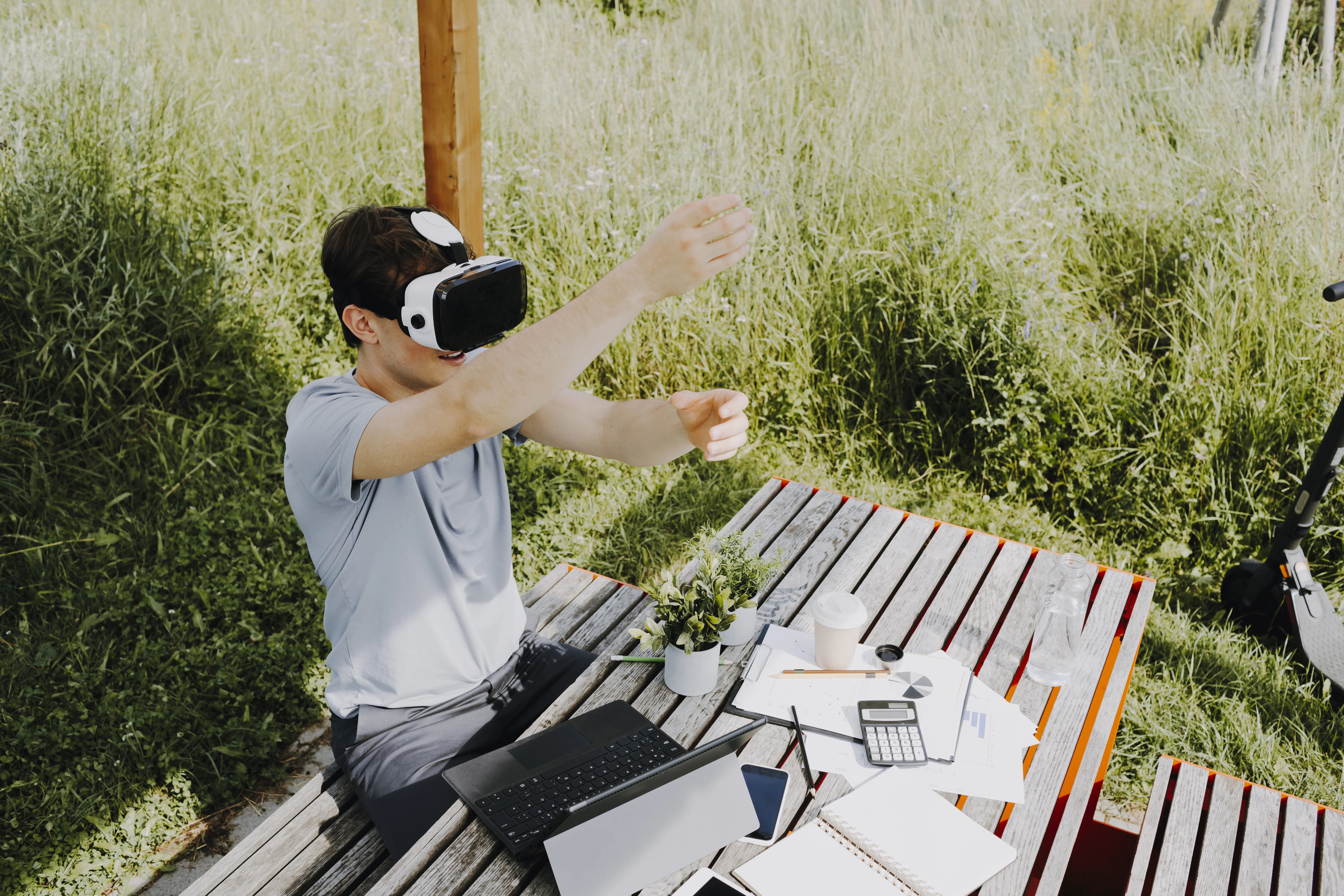 Ein junger Mann sitzt auf einer Holzbank mit aufgesetzter Virtual-Reality-Brille; auf dem Tisch liegen Arbeitsunterlagen, ein Notizbuch und Pflanzen, und im Hintergrund steht ein E-Scooter. Person